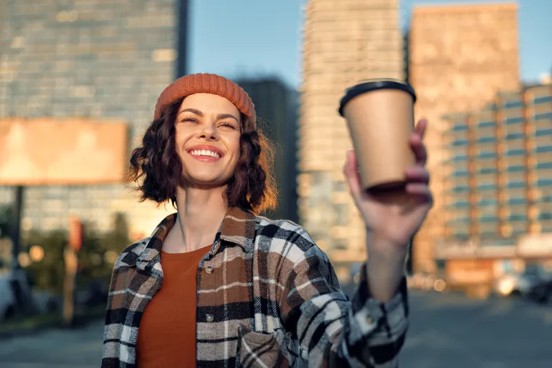 Woman coffee smile city beanie sunlight takeaway portrait captured in candid lifestyle moment with golden hour glow, authenticity and emotional storytelling for mindful living and warmth.