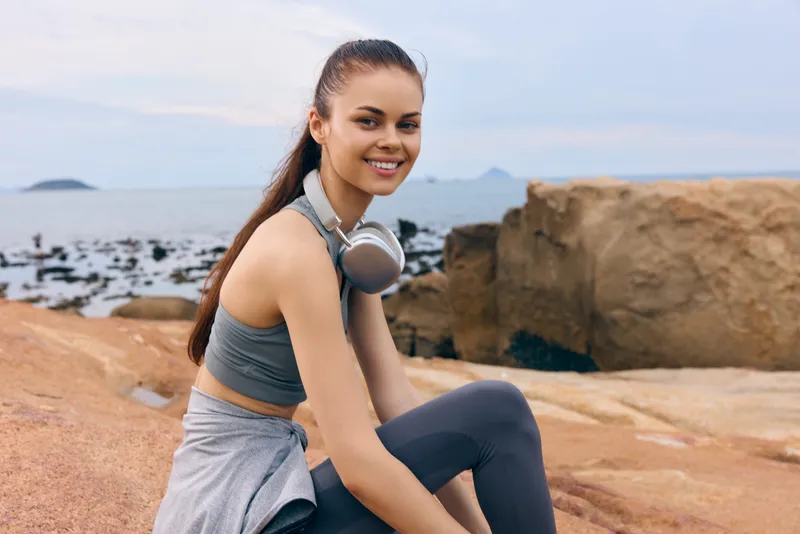 Young woman exercising outdoors on rocky beach, wearing sportswear, smiling confidently, with ocean in the background, showcasing an active lifestyle
