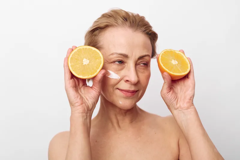 Mature woman applying facial cream holds two orange halves near her face, demonstrating natural skincare and healthy lifestyle concept on white background.