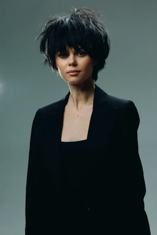 Young woman with short black hair wearing a black outfit in a minimalist dark studio setting.