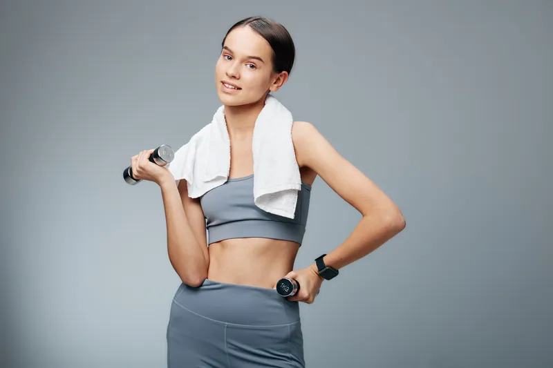 Woman fitness, sport and workout with dumbbells, wearing a gray athletic set and a towel around her neck, posing in a clean studio for health and exercise motivation.