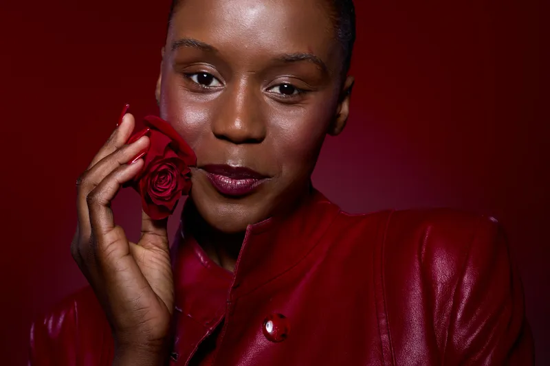 Close-up of a dark skinned African woman holding a red rose near her face. She wears a red leather jacket with a confident expression on a deep red background. Beauty and elegance concept.