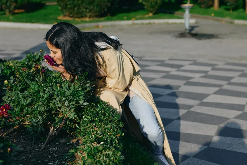 Flowers surround a woman as she leans toward a flowering bush enjoying a soft scent in a sunlit garden, casual coat, serene moment of nature and calm exploration