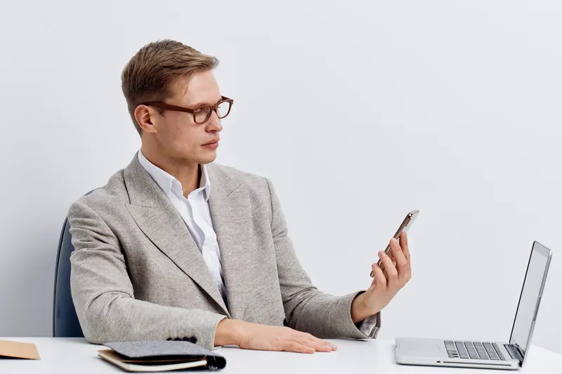 Professional young businessman with glasses wearing grey suit and white shirt looking at smartphone, sitting at desk with open laptop and notepad, isolated on plain white background, studio portrait. 