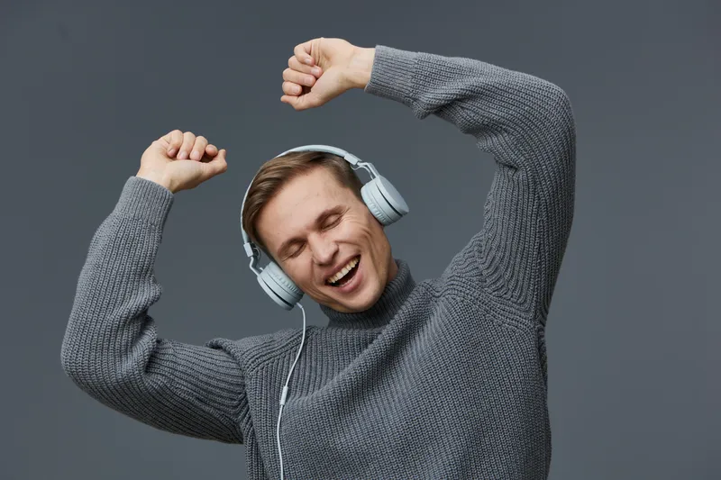 happy young man in headphones listening music with closed eyes dancing raised hands wearing gray knit sweater isolated on gray background digital detox concept
