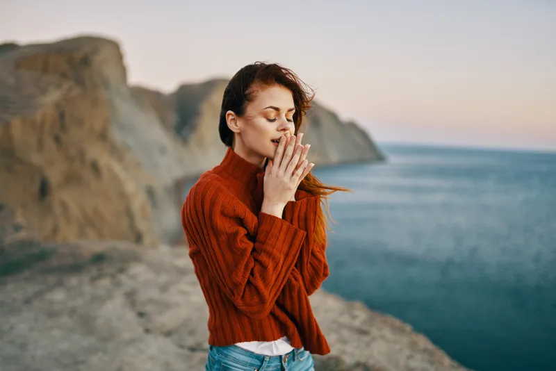 Рыжеволосая девушка в красном свитере на скалистом берегу моря