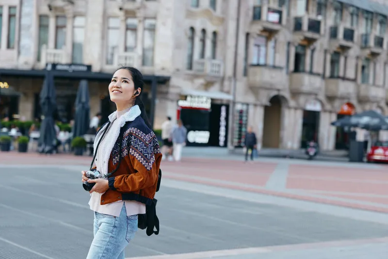 Female street photographer in a patterned jacket and scarf walks across an urban square, camera in hand, sunlight, historic buildings in the background during daytime.