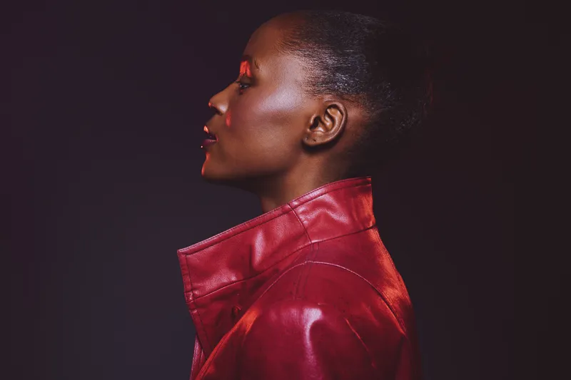 Profile of a dark skinned African woman wearing a red leather jacket and red eye makeup. Studio portrait with dramatic lighting on a dark background. Concept of fashion and beauty.