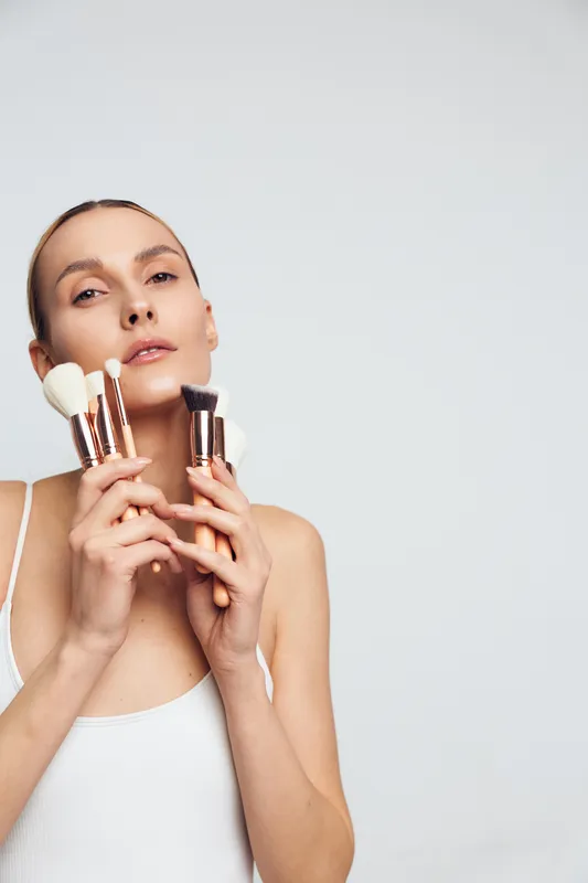 Young woman with short blonde hair holds makeup brushes near her face while posing confidently against a plain light background in a white tank top.