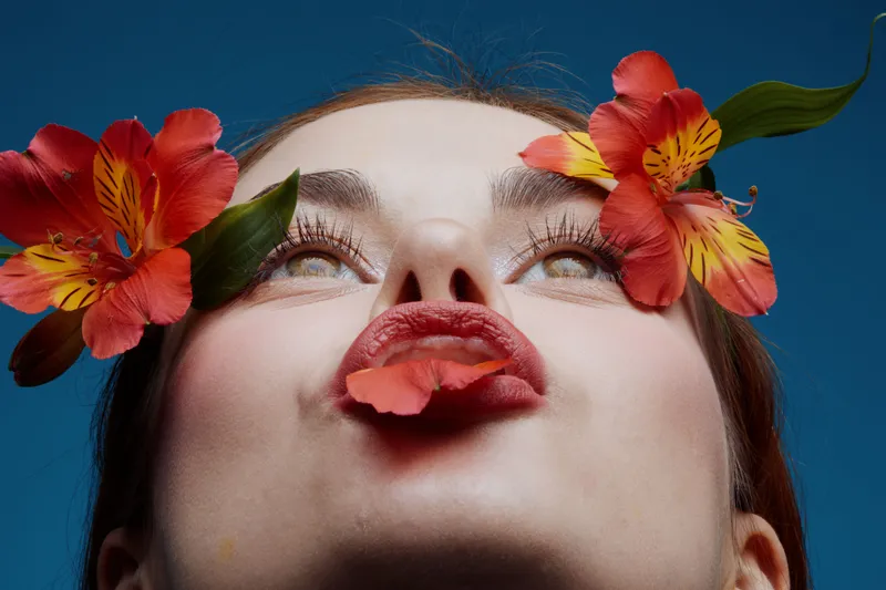 Young woman with red lips and colorful flowers, expressing joy, against a vibrant blue background, focusing on beauty and nature in spring