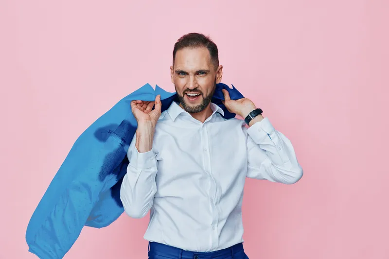 A professional man in business attire stands against a solid pink background, draping a blue jacket over his shoulder to convey confident energy and a bright mood.
