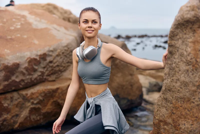 Young woman in activewear smiling outdoors near the ocean with headphones on, capturing a joyful and healthy lifestyle