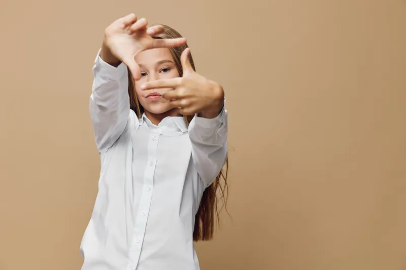 Girl posing with hands framing face in creative gesture, wearing white shirt, standing against a beige background in studio for portrait or expression concept.