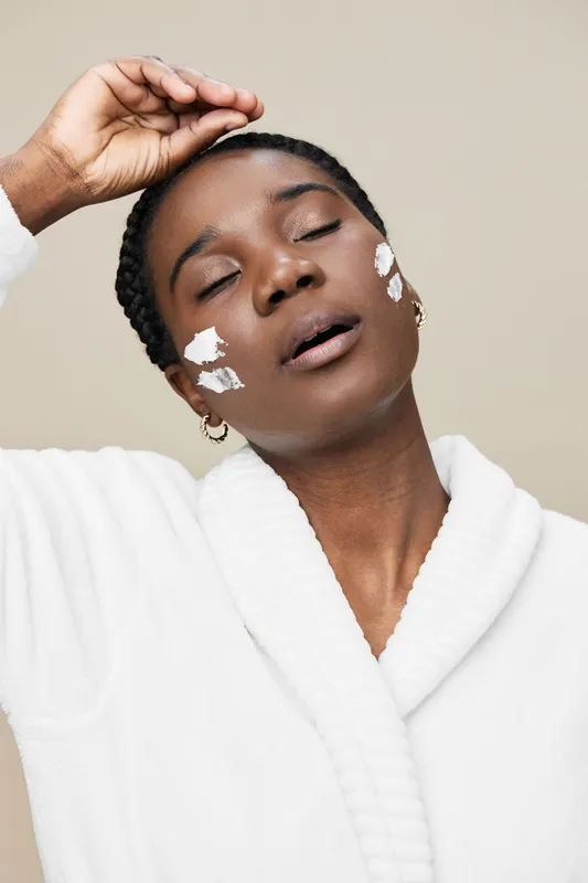 Relaxed African woman enjoying a skincare treatment with white cream on her face, showcasing a serene expression against a soft beige background