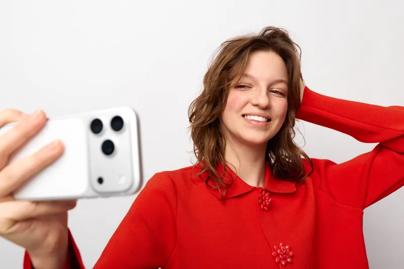 Smiling young woman in red sweater takes selfie with smartphone on white background. Casual lifestyle, happiness, and modern technology concept with natural expression and relaxed pose.