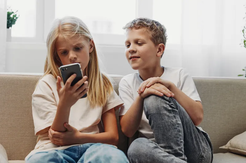 Two children sit on sofa, girl focused on smartphone while boy looks interested. Concept of childhood, technology, communication, and curiosity.