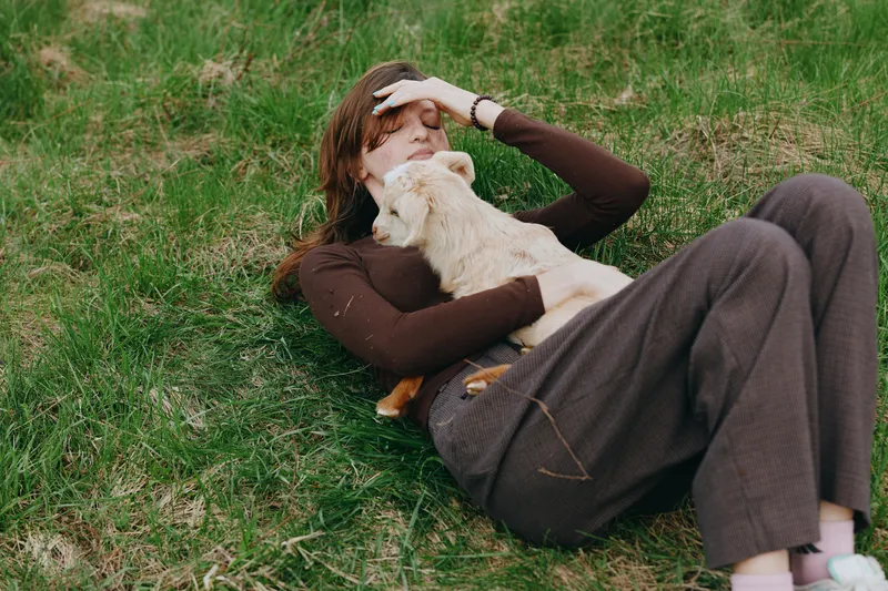 Young woman rests on grass outdoors, cuddling a friendly goat kid, expressing calm affection and a simple lifestyle moment, cozy brown clothes create a warm, intimate countryside scene.