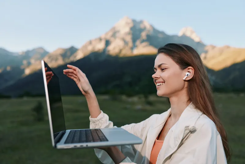 A cheerful professional woman uses a laptop outdoors, smiling as she leads a virtual meeting in a bright field with mountains in the distance, creating a positive productive mood.