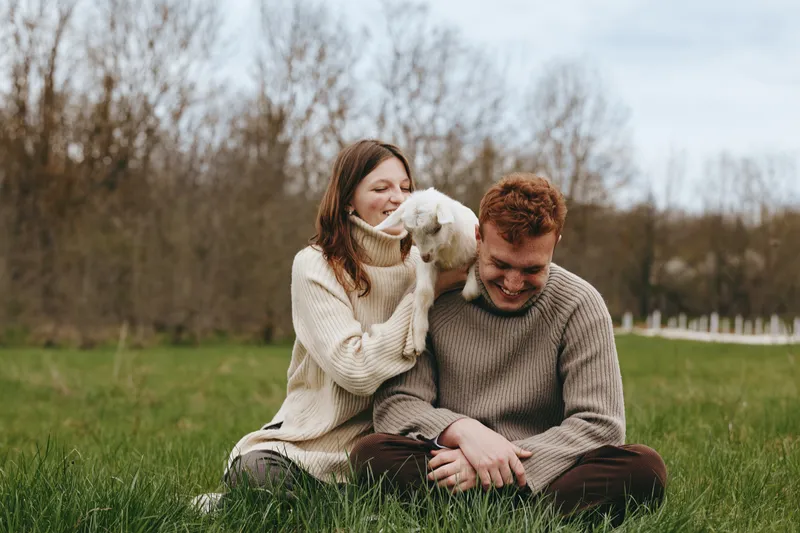 Happy couple shares a tender moment with a playful goat kid in an open meadow, sitting on grass, cozy sweaters, warm smiles, peaceful countryside vibe, candid lifestyle scene.