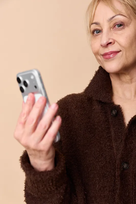 Elderly woman portrait holding a smartphone, calm smile in a dark coat on a beige studio background, using technology for communication on a touchscreen for everyday life.