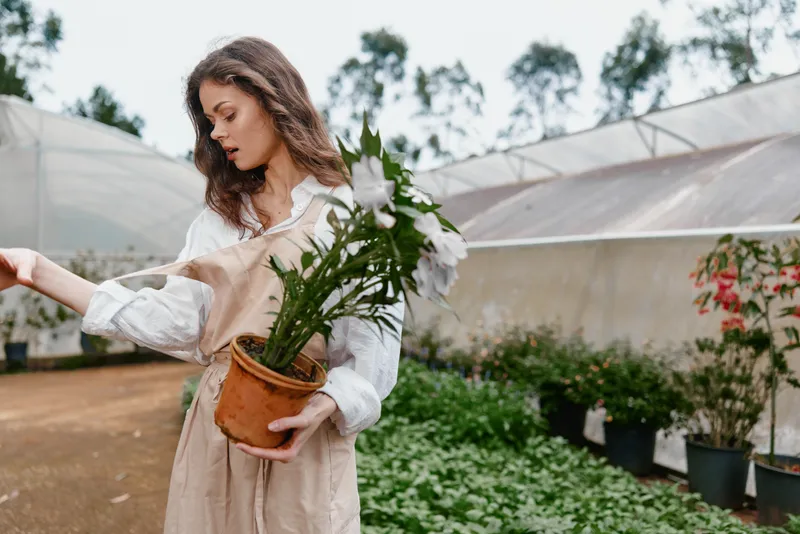 Young woman in light clothing holding a potted plant in a greenhouse, showcasing a connection with nature and gardening passion, ideal for seasonal themes of growth and renewal
