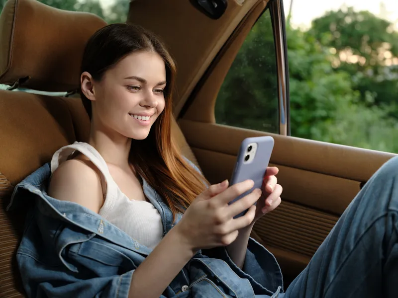 Young woman enjoying her smartphone in a car, wearing denim jacket, smiling, casual outfit, lush green background, summer vibe, relaxation concept.