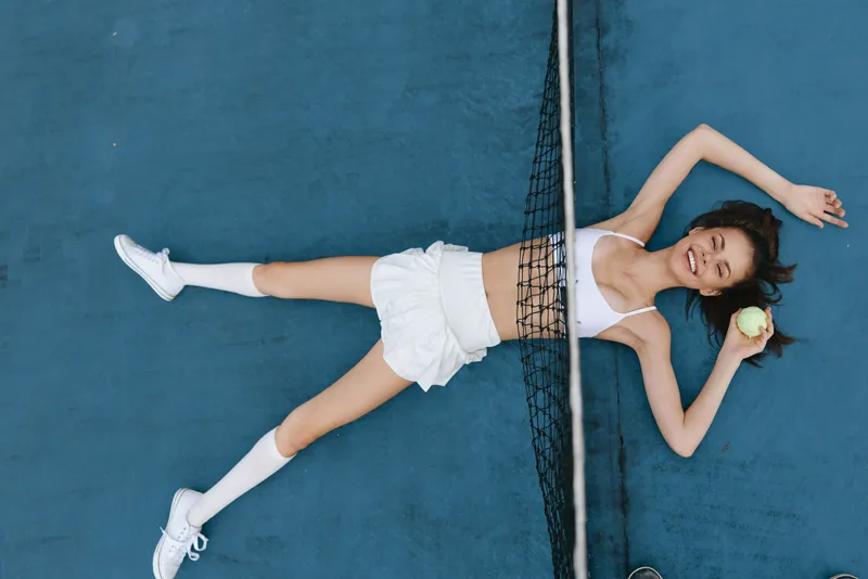 Young Asian woman in white athletic wear lying on a tennis court, holding a tennis ball, showcasing a playful and relaxed attitude on a sunny day