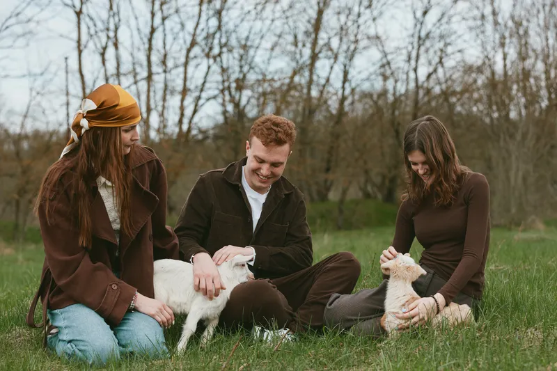 A lifestyle scene outdoors with animals, goats, and kids as three friends relax in a sunny field, sharing smiles and gentle pets in a wholesome countryside moment.