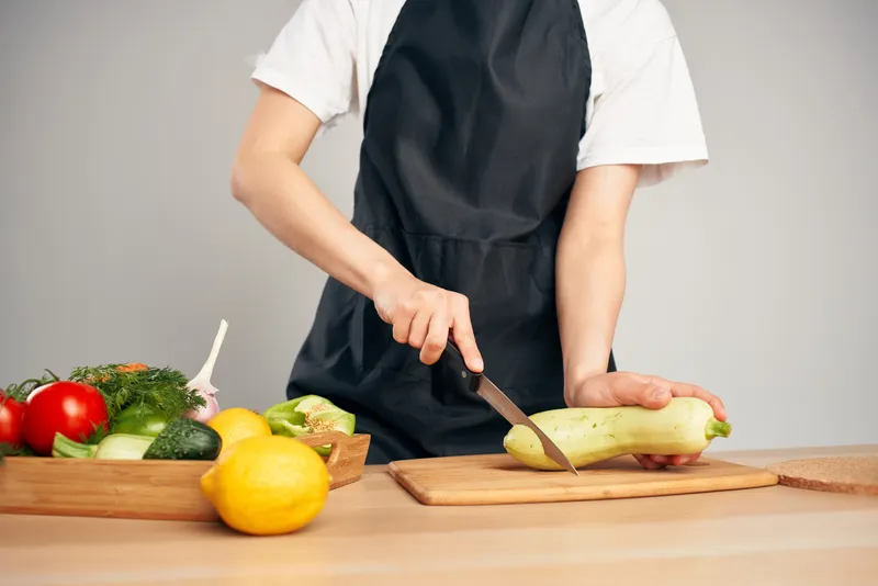 Женщина в черном фартуке нарезает овощи на кухне, готовя еду