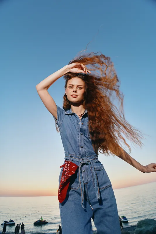 young woman with long curly hair wearing denim outfit and red bandana by the sea during sunset outdoor casual style