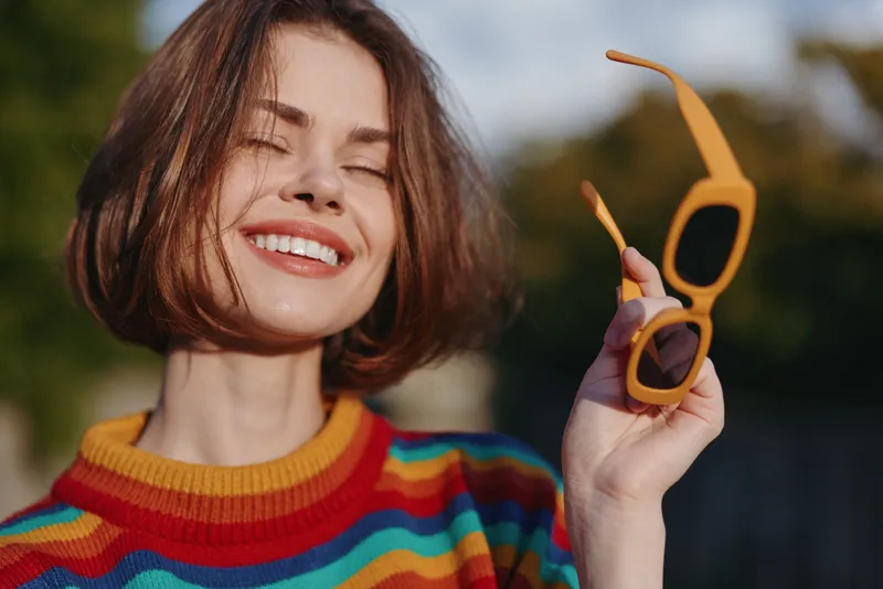 Woman smiling in colorful striped sweater, short hair portrait holding yellow sunglasses outdoors, eyes closed and joyful, casual outfit, lifestyle fashion for travel and leisure.
