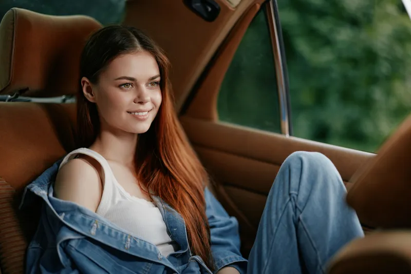 Smiling young woman with long red hair wearing a denim jacket and white tank top, relaxing inside a car, against a natural green background.