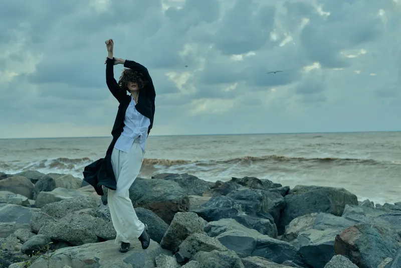 Woman leaps on rocky shore beside the sea, wind catching her jacket, rugged coastline and dramatic clouds creating a dynamic outdoor fashion moment.