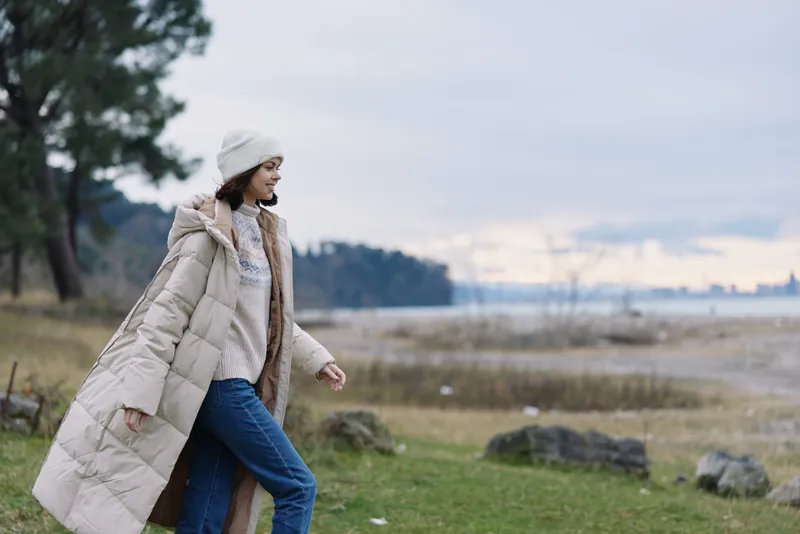 Woman walking outdoors in a long beige coat, knitted hat and jeans along a windy coastal park with sea, trees and rocky shoreline.