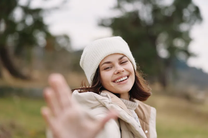 Smiling woman outdoors with stop gesture, reaching hand in a friendly portrait amid nature, casual clothing and winter wear in daylight.