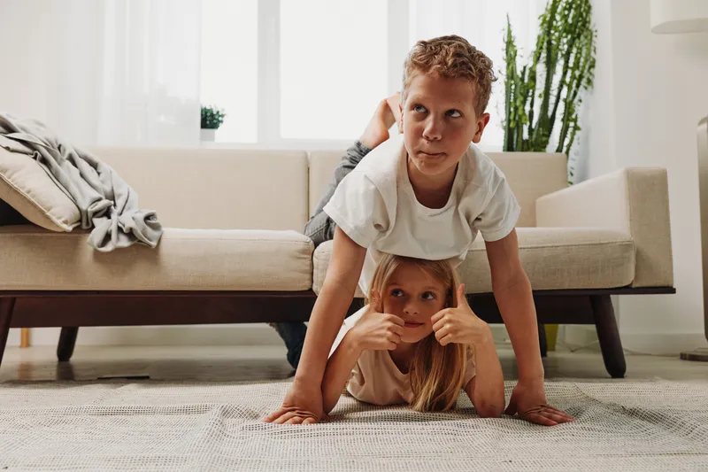Two playful children in casual clothes perform a balancing pose on a carpet in a bright living room with modern furniture and natural light. Concept of family fun and sibling bonding.