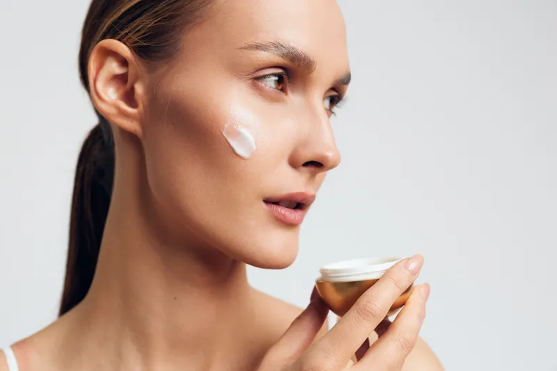 Young woman with clear skin applying white face cream on cheek, holding jar. Skincare and beauty concept with soft natural lighting on a white background, close-up portrait of healthy skin care.