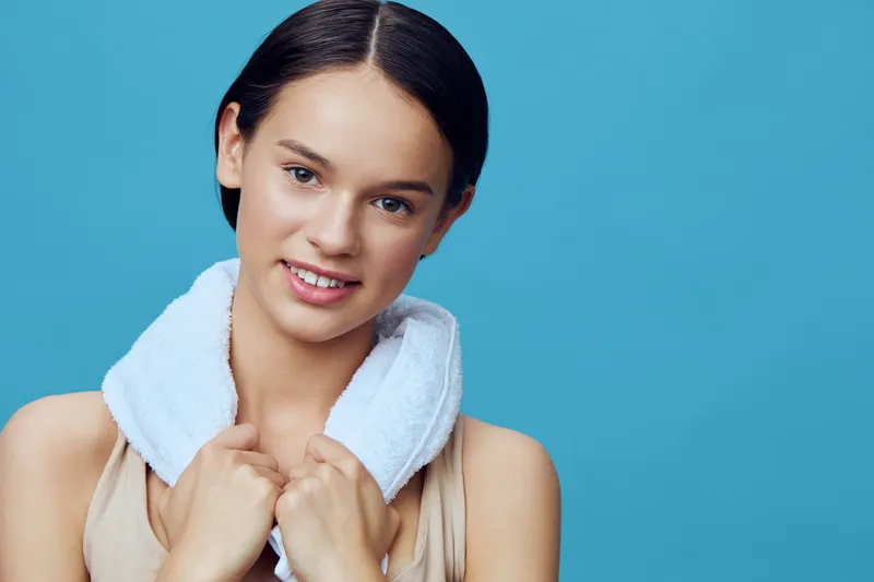 Young female fitness model with a towel on her shoulders, smiling against a turquoise background, exuding health and vitality in a stylish sports outfit
