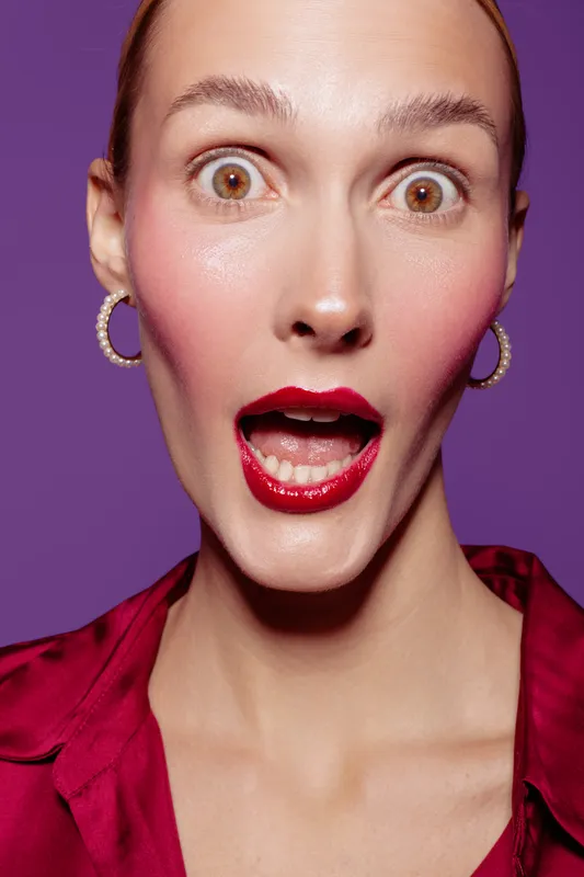 Surprised woman with wide open eyes and mouth wears red lipstick and earrings. Close-up portrait on purple background. Concept of shock, amazement, and emotional reaction.