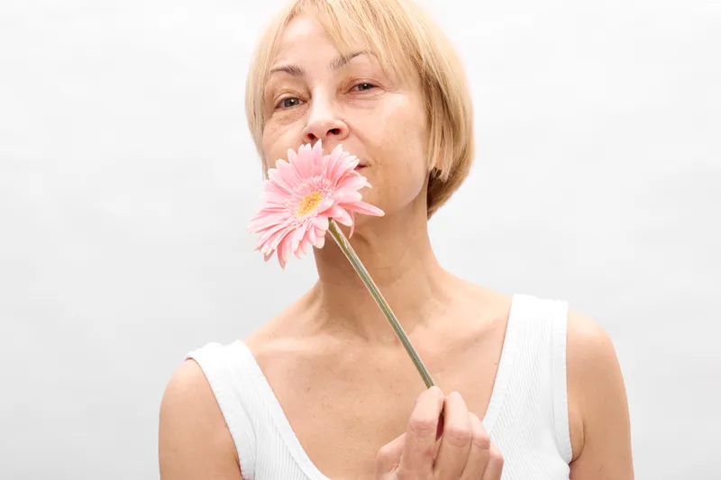 Woman beauty portrait holding a pink flower near her lips, smiling softly, practicing skincare and wellness. White studio background, spring mood, soft light.