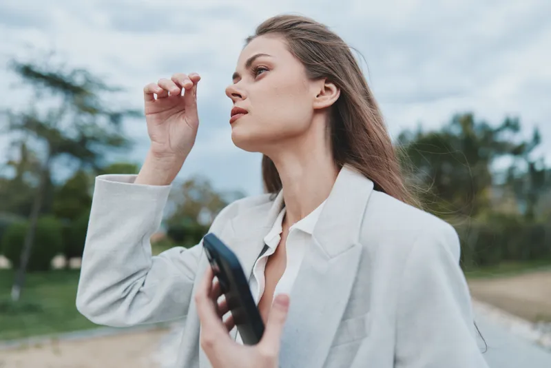 Woman in white blazer outdoors holding phone displaying thoughtful expression in natural background, business concept portrait