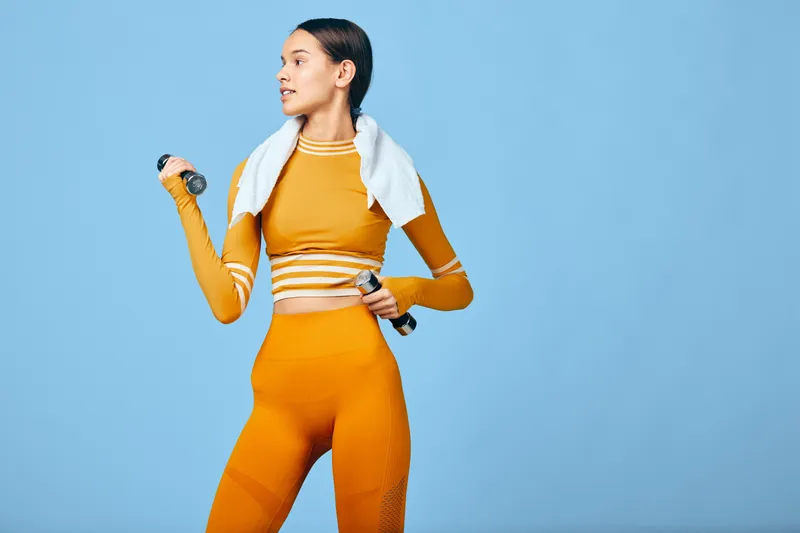 Fitness woman in orange athletic wear with a towel over her shoulders, holding a dumbbell during a workout against a blue backdrop, showcasing sport, strength and dedication.