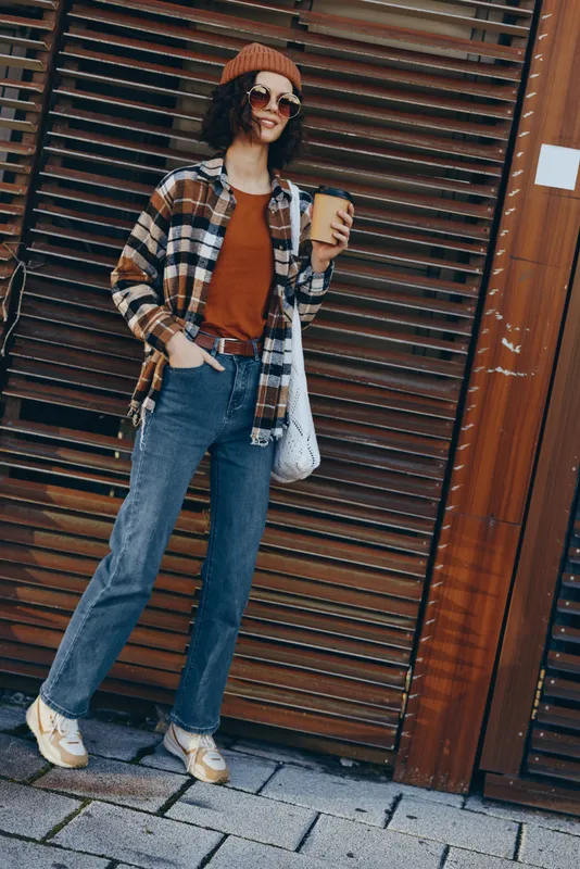 Woman holding coffee on autumn street in casual beanie and plaid jacket with sunglasses and smile, urban candid lifestyle scene emphasizing authenticity, mindful living and golden hour glow.