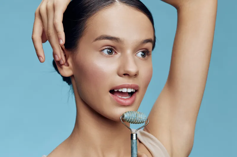 Young woman using a facial roller against a vibrant blue background, showcasing radiant skin and a joyful expression, perfect for beauty and skincare themes