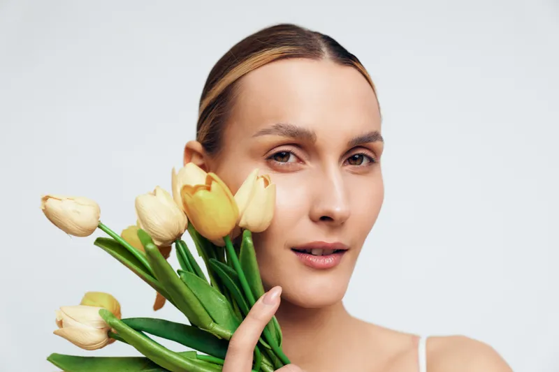 Young woman holding yellow tulips close to her face, natural makeup, soft lighting, calm expression, minimalistic background, fresh flowers, spring mood, gentle atmosphere