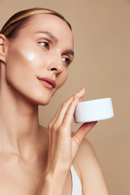 Young woman with clear skin applying white cream on face, holding cosmetic jar, skincare routine, natural beauty, neutral background, soft lighting, healthy skin care concept