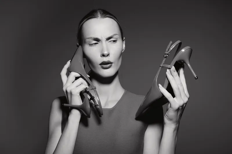 Confused Caucasian woman with pale skin holds high heel shoes, one near ear, in studio with dark background. Concept for fashion dilemma, choice and style.