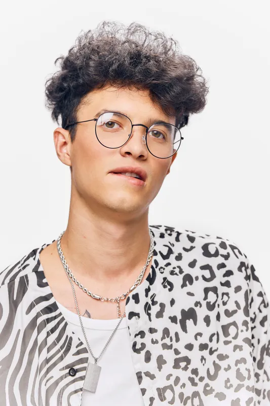 young man wearing glasses and a patterned shirt, with curly hair and a contemplative expression against a plain white background