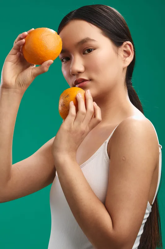 asian woman holds oranges against a vibrant green backdrop, close to her face with a calm expression, sunlight highlights her skin, creating a fresh, modern still life portrait.