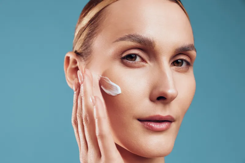 Close-up of a young woman with brunette hair applying white face cream on her cheek against a blue background, expressing calmness and skincare routine.